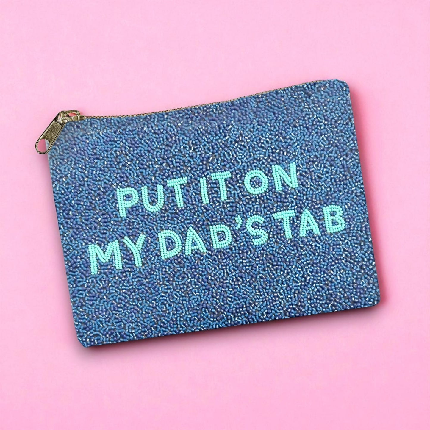 Glittery beaded zipper pouch with playful “Put It On My Dad’s Tab” text, laid flat against a pink backdrop.