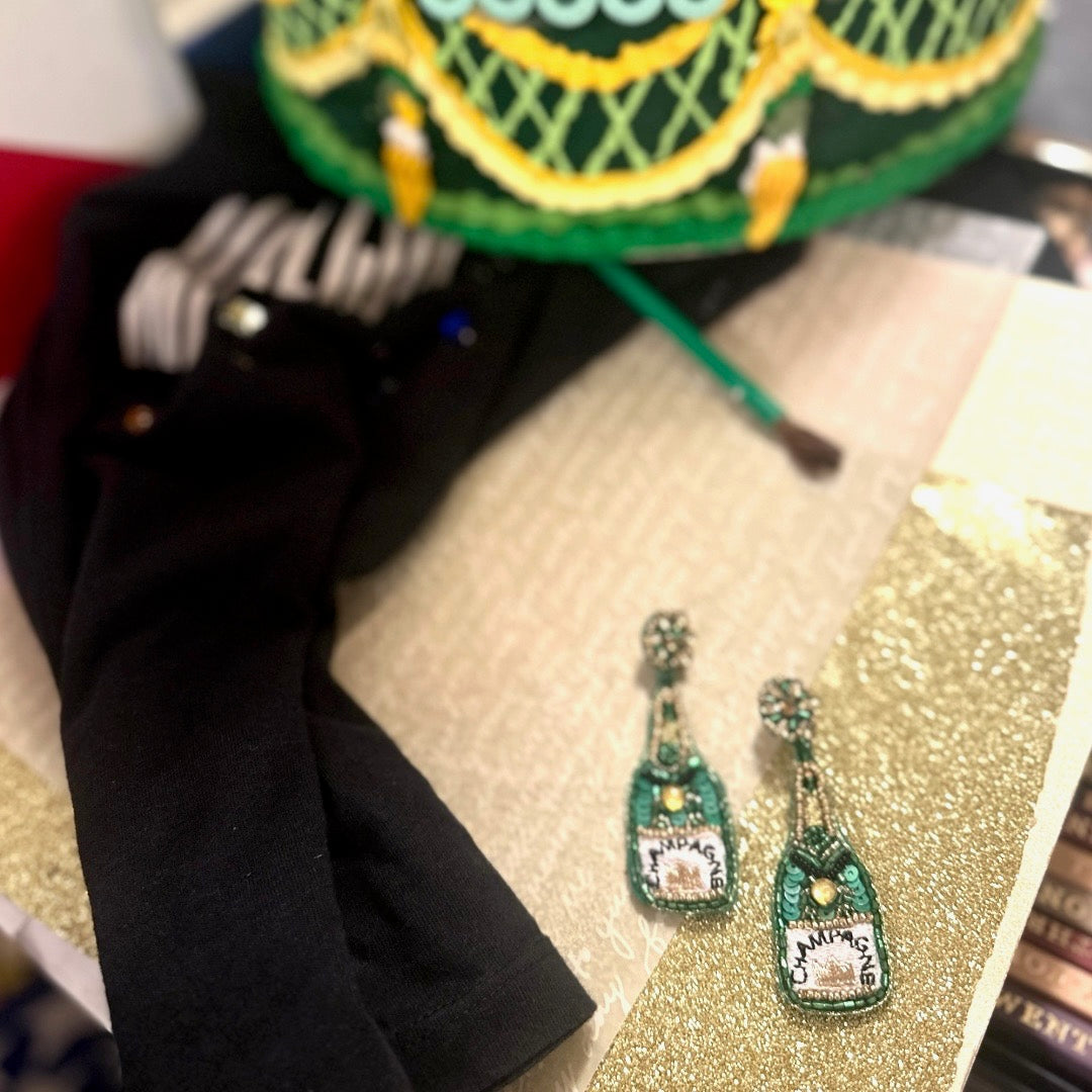 Beaded statement earrings shaped like green champagne bottles with rhinestone tops, placed on gold and cream shimmer fabric next to a green embroidered hat and black fabric.