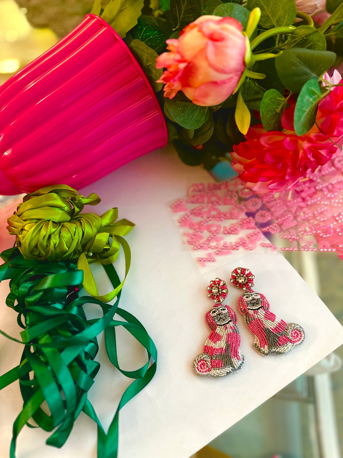 Whimsical pink and silver dog-shaped beaded earrings styled on white paper with ribbon, faux flowers, and a hot pink flower pot spilling a vibrant bouquet.