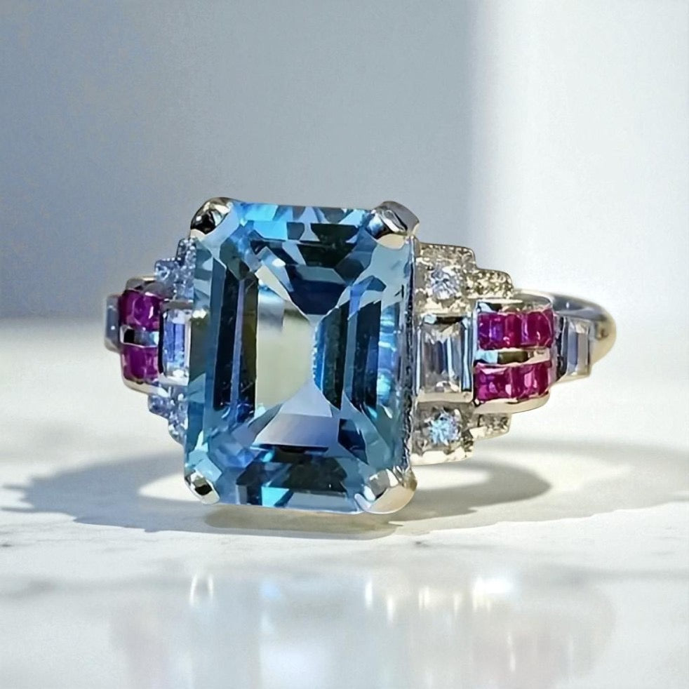 Emerald-cut blue gemstone ring set in silver with sparkling diamond and ruby accent stones on each side, displayed on a white marble surface with natural lighting.