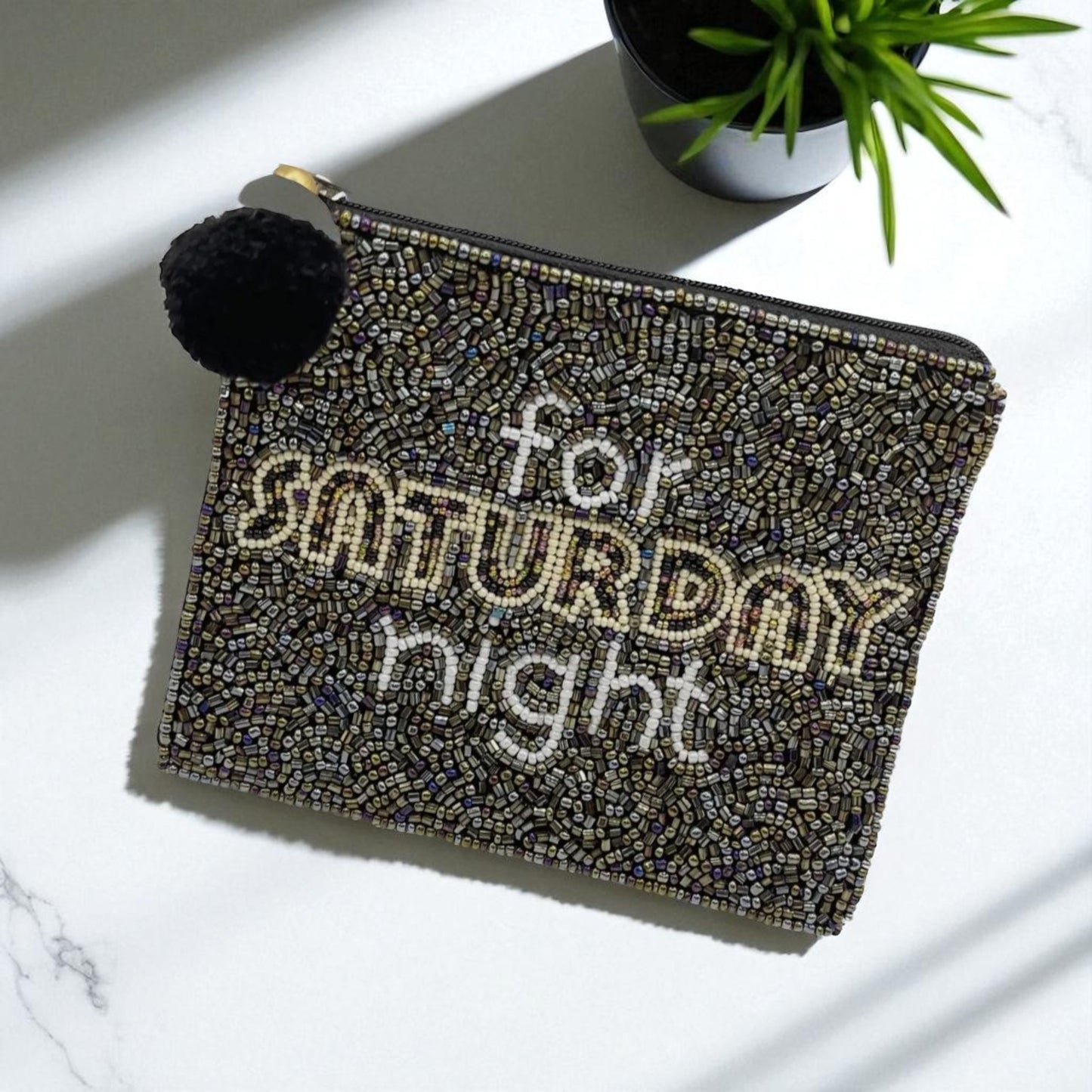 Beaded clutch bag with a black and gold mosaic pattern, featuring the phrase “for SATURDAY night” in white and gold lettering, topped with a black pom-pom zipper pull, placed on a white marble surface next to a potted plant.