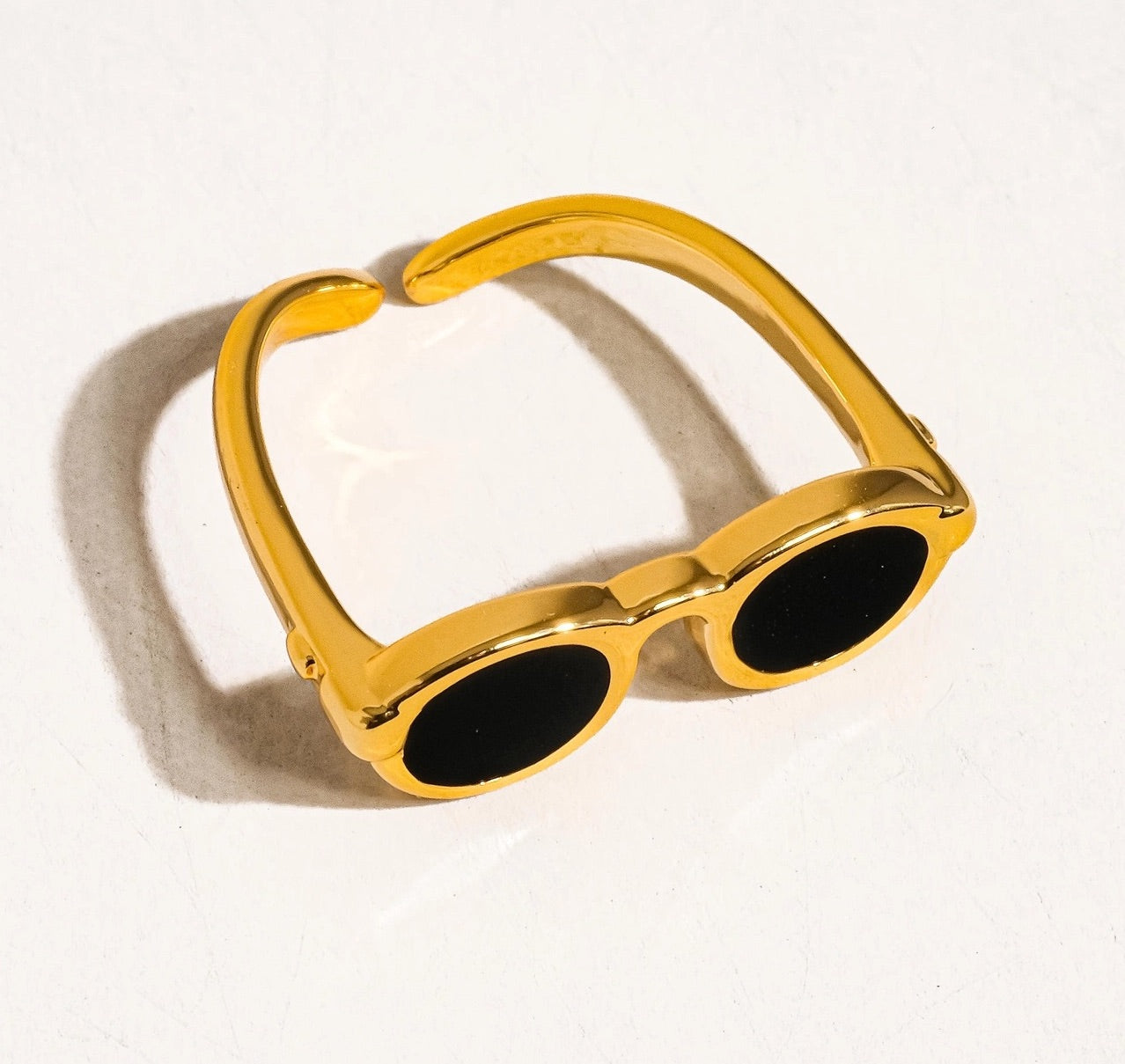 Gold ring shaped like a pair of sunglasses, shown from a side angle on a white background, casting a soft shadow.