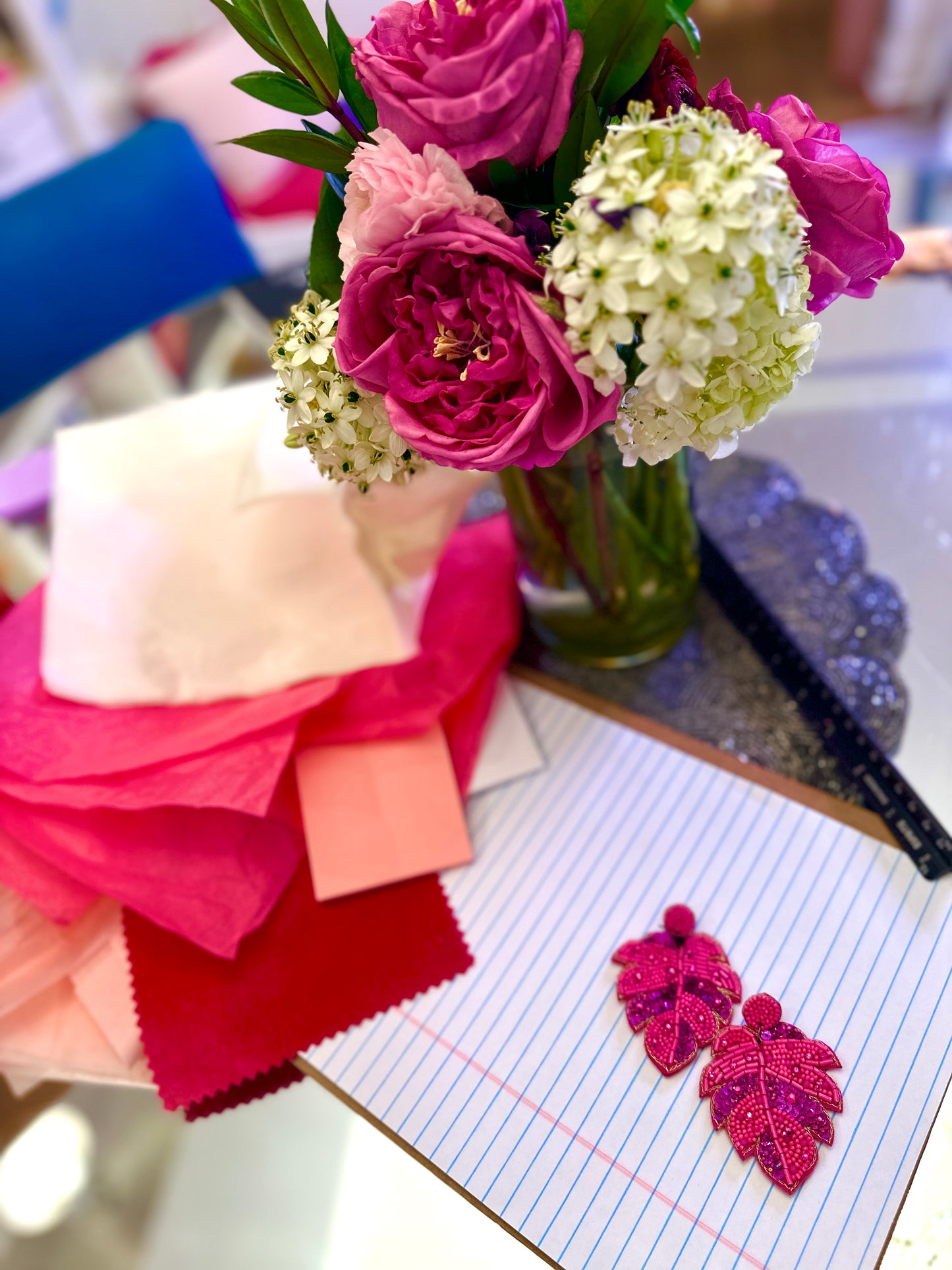 Bright pink beaded leaf-shaped earrings styled with matching fabric swatches and a bouquet of fresh flowers in shades of pink and white.