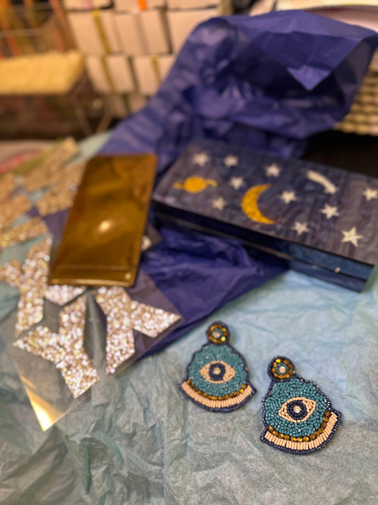 Mystical Misfit Blue and Bronze Earrings with evil eye motif displayed on soft tissue paper alongside a celestial-themed clutch and glittering star stickers. Vibrant and whimsical styling for a mystical vibe.