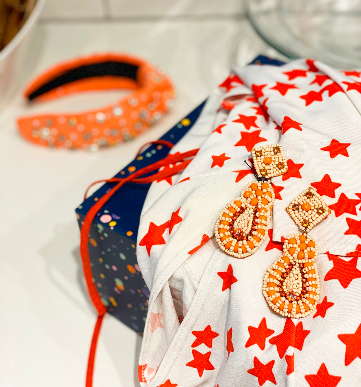 Bright and playful product styling featuring vibrant orange and cream beaded earrings paired with a starry red and white print and bold accessories—perfect for celebrating color and creativity in your look.