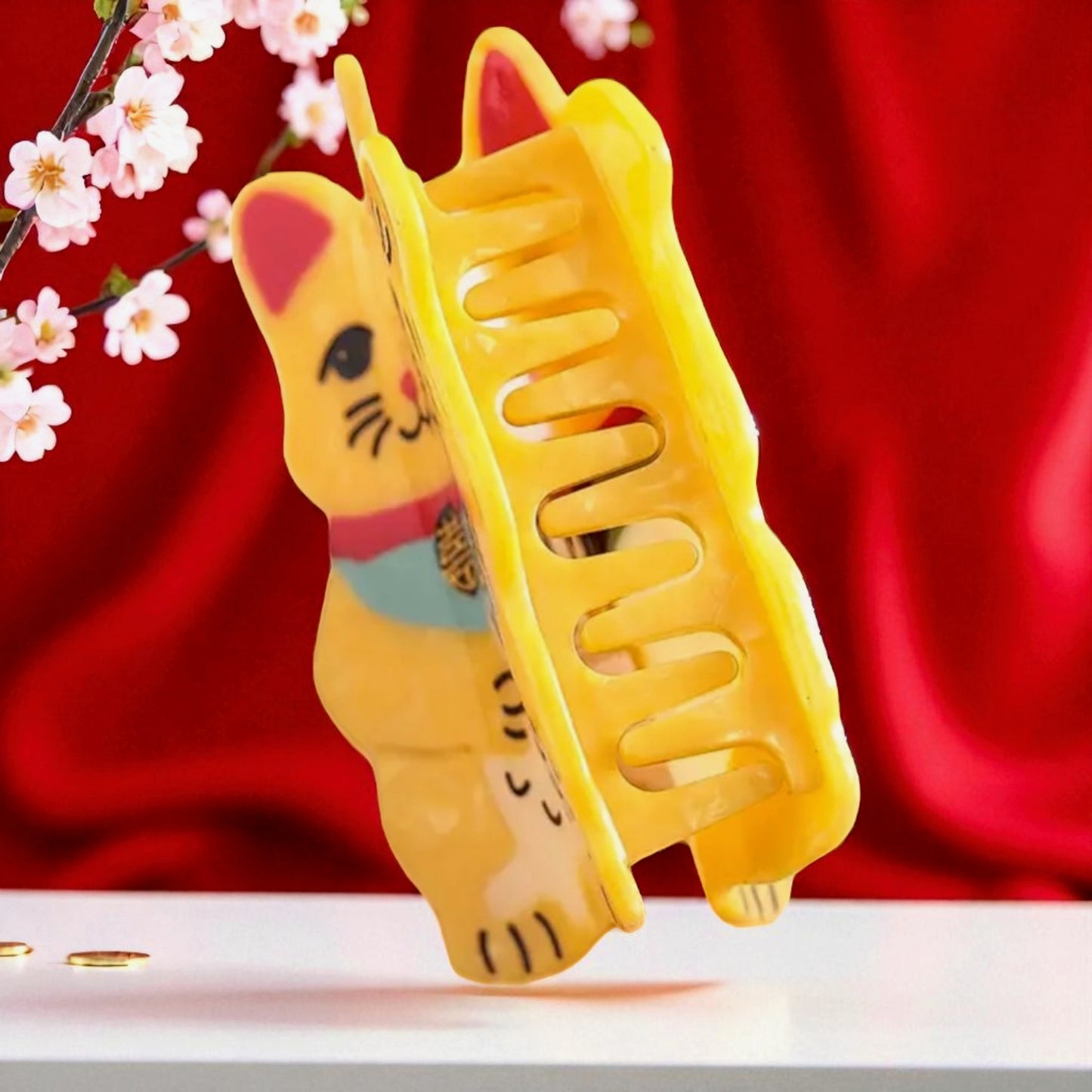 Side profile of a Maneki Neko-shaped yellow hair claw clip displayed upright on a white surface with cherry blossoms, red background, and a festive Lunar New Year theme.