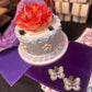 Pair of iridescent purple and gold butterfly-shaped sequin earrings displayed on deep purple velvet next to a pastel cake with a bright flower topper.