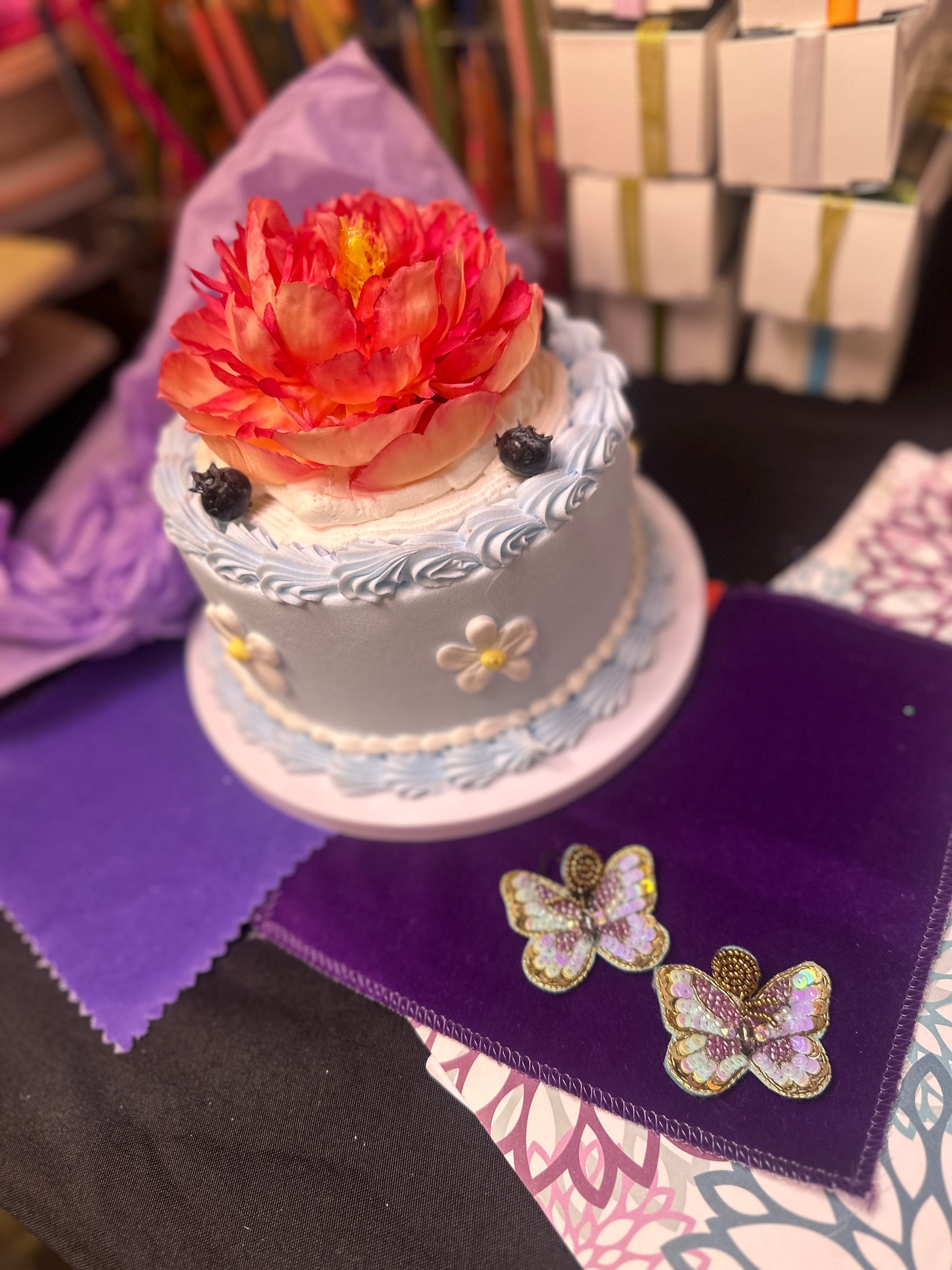 Pair of iridescent purple and gold butterfly-shaped sequin earrings displayed on deep purple velvet next to a pastel cake with a bright flower topper.