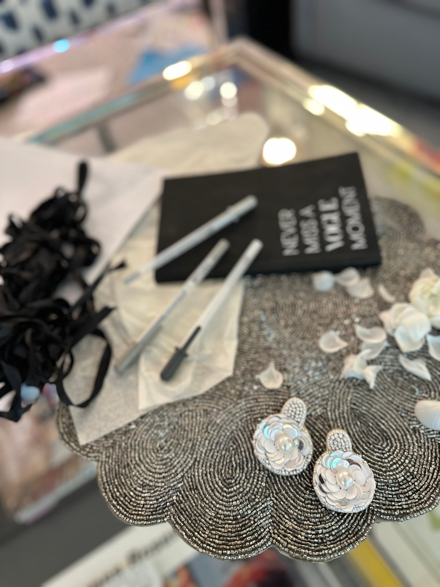 Iridescent white floral earrings with sequin petals and pearl centers, placed on a silver beaded placemat alongside white petals, black ribbon, and a notebook.