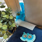 Beaded fringe earrings featuring a blue evil-eye design, styled on a navy cushion with greenery and soft window light.