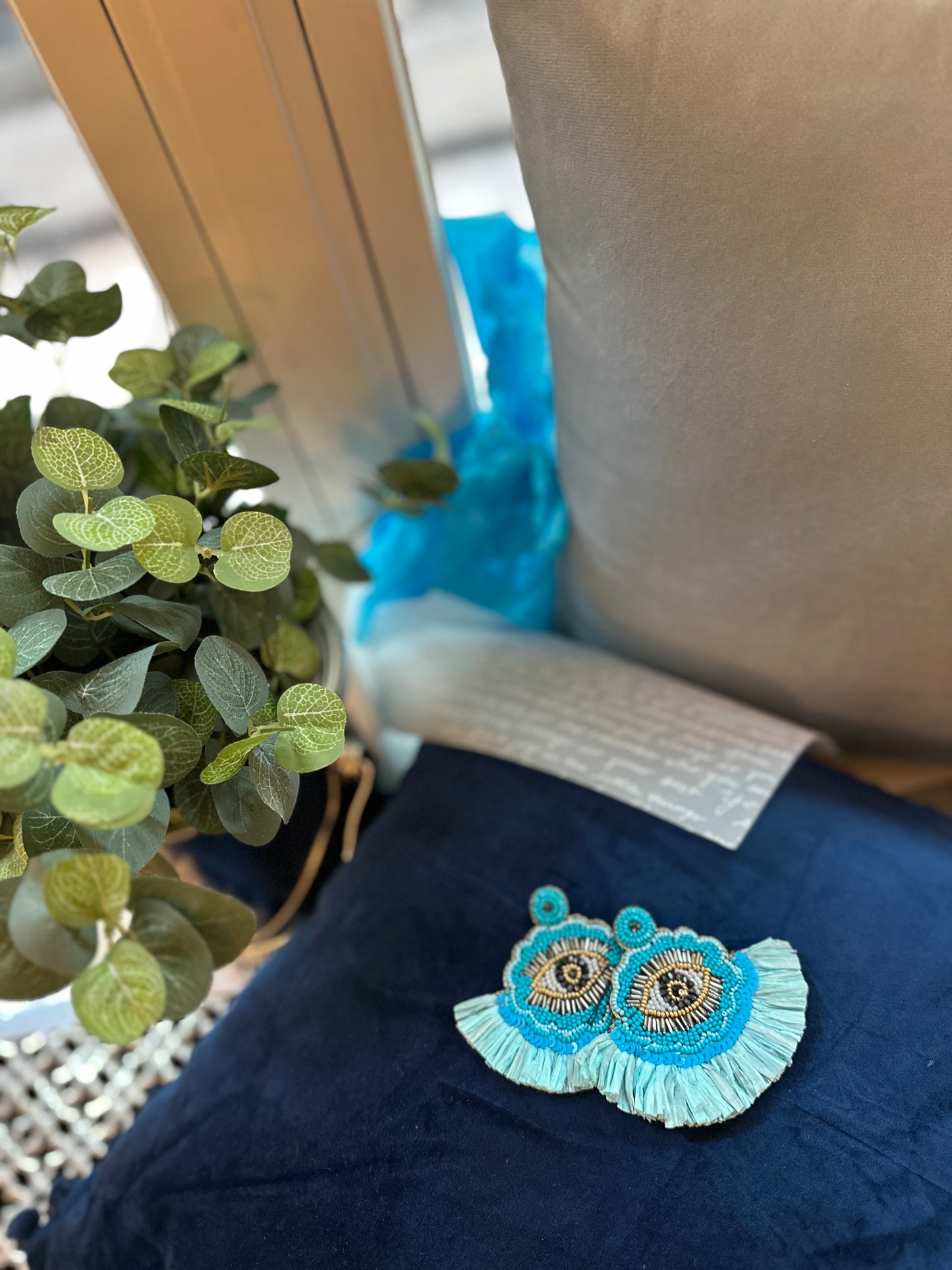 Beaded fringe earrings featuring a blue evil-eye design, styled on a navy cushion with greenery and soft window light.
