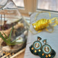 A pair of elegant green statement earrings with beaded centers and layered dark green tassels, accented by gold discs, displayed on a soft gray velvet pillow near yellow ribbon and a glass terrarium with plants.