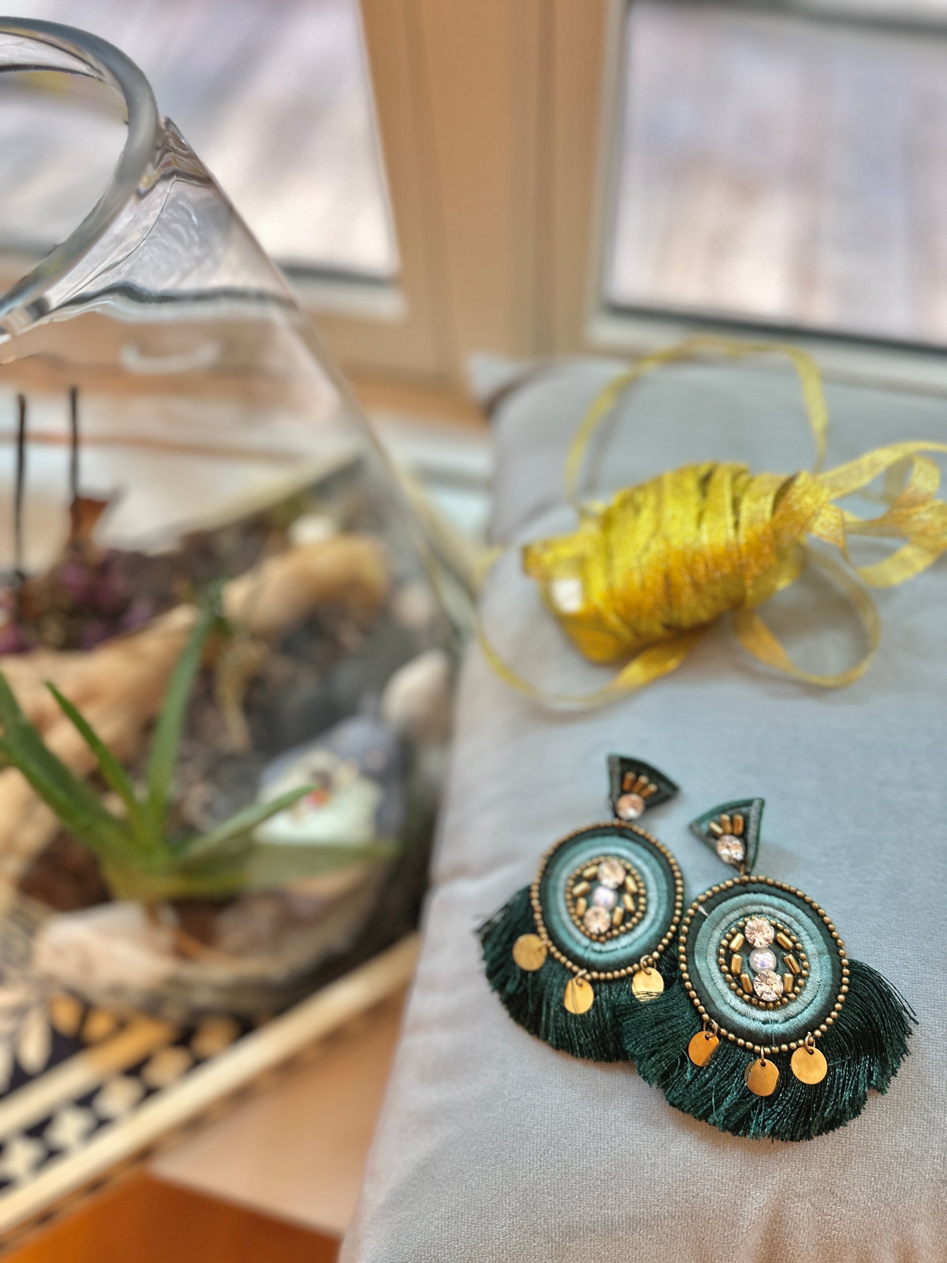 A pair of elegant green statement earrings with beaded centers and layered dark green tassels, accented by gold discs, displayed on a soft gray velvet pillow near yellow ribbon and a glass terrarium with plants.