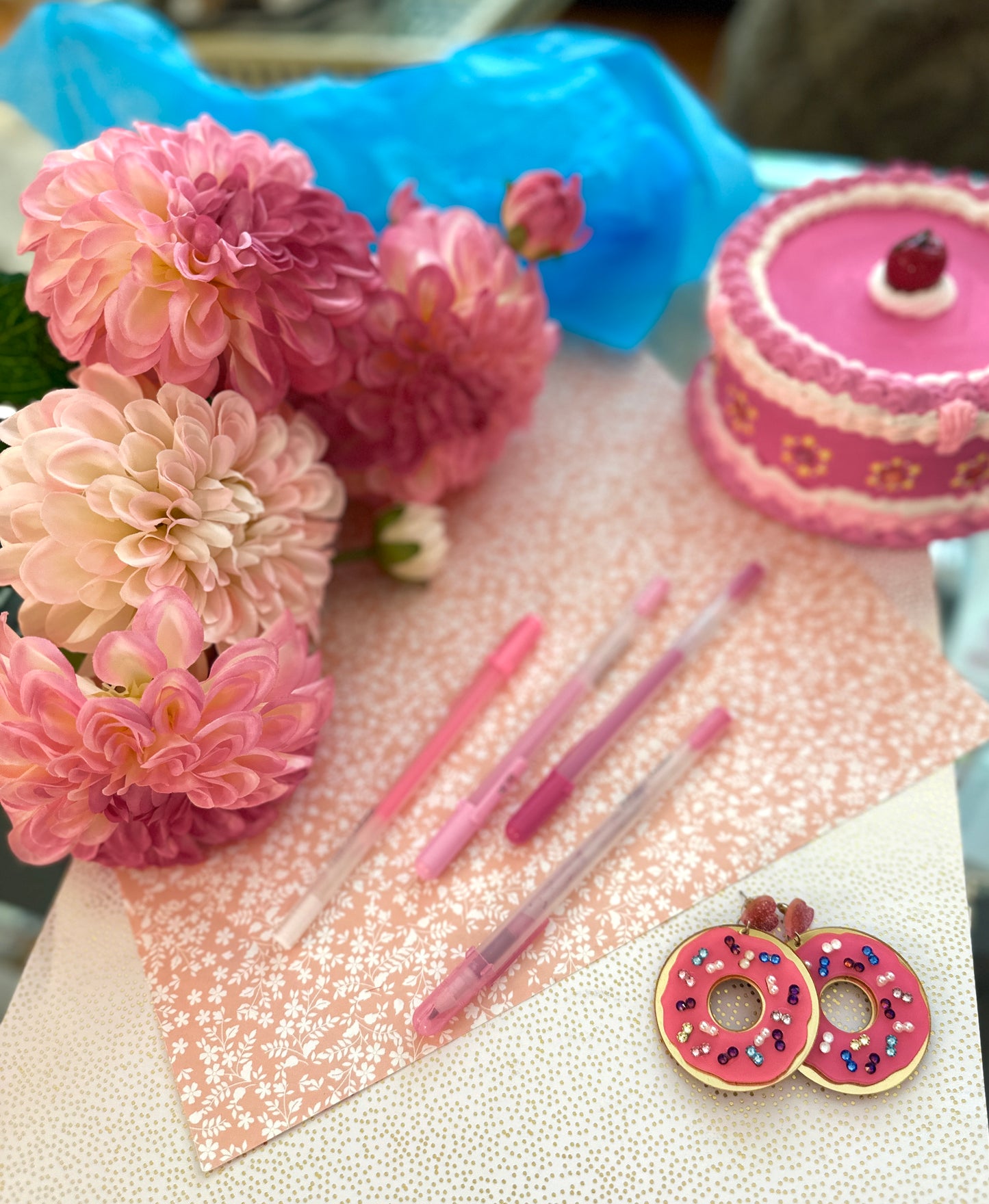Playful pink donut-shaped earrings with multicolor sequins, styled with floral stationery, pink pens, and a mini cake for a whimsical look.