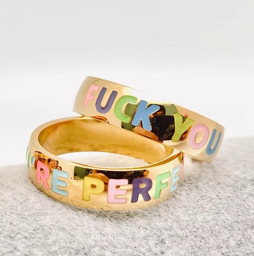 Stack of two gold rings with rainbow enamel lettering, one saying "FUCK YOU" and the other "YOU'RE PERFECT", cheeky and colorful jewelry set
