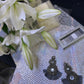 Sparkling gunmetal black crystal earrings with layered beading and black fringe, displayed on iridescent paper next to blooming white lilies and silver eyeshadow palette.