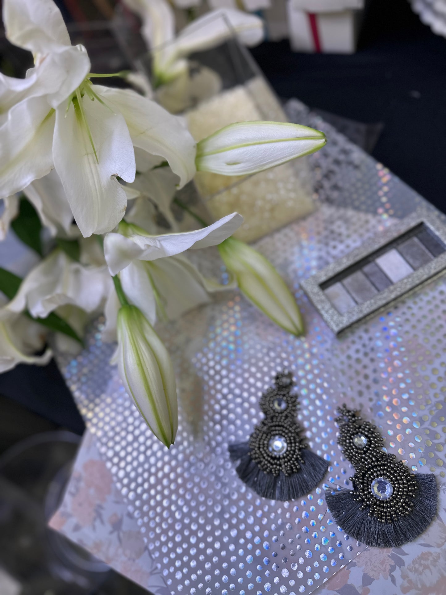 Sparkling gunmetal black crystal earrings with layered beading and black fringe, displayed on iridescent paper next to blooming white lilies and silver eyeshadow palette.