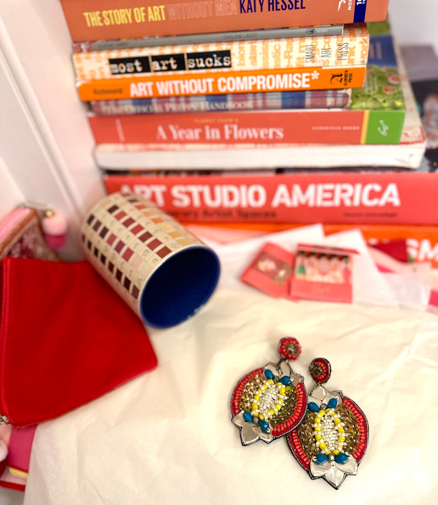 Colorful galactic-inspired beaded earrings styled against a backdrop of stacked art books, red accessories, and paint swatches.