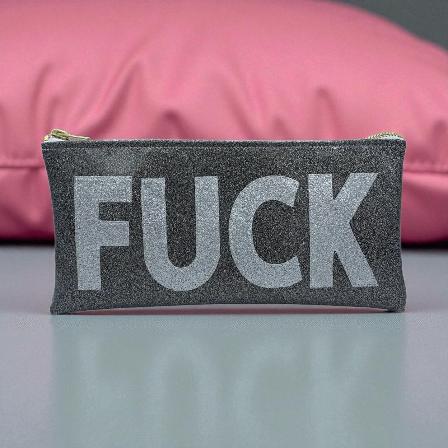 Small black glitter pouch with bold silver “FUCK” lettering, styled in front of a pink pillow.