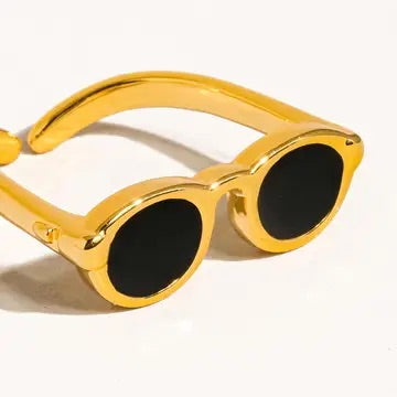 Angled close-up of a gold sunglasses-shaped ring, highlighting the glossy finish and black lens detailing on a cream background.