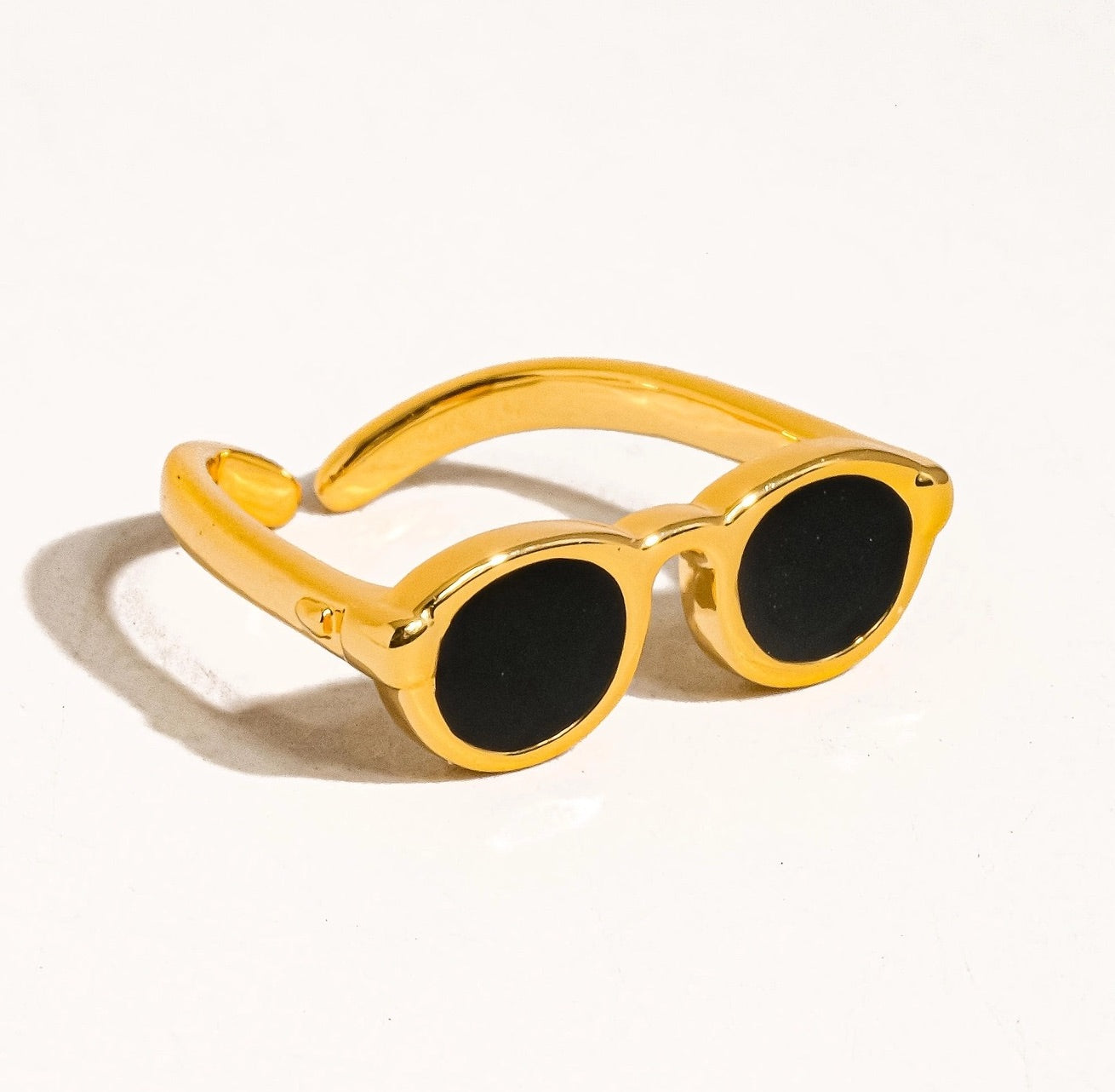 Flat lay of a bold gold ring modeled after sunglasses, placed on a white surface with visible shadowing around the edges.