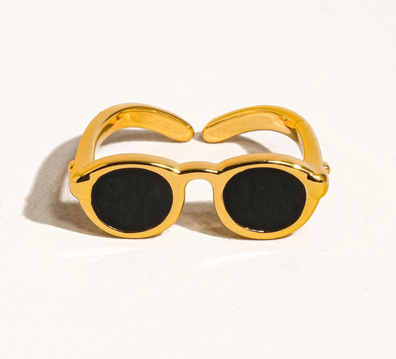 Front-facing view of a shiny gold ring designed to resemble sunglasses, with round black lenses and open-ended arms.