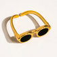 Gold ring shaped like a pair of sunglasses, shown from a side angle on a white background, casting a soft shadow.