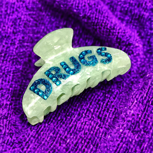 "DRUGS" hair clip with green acetate base and blue rhinestone lettering.