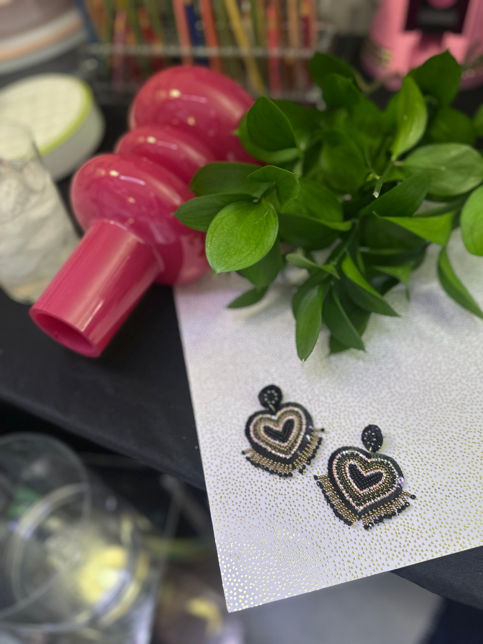 Black and gold beaded heart earrings with fringe, placed on metallic dotted paper beside a lush green plant and bold pink vase.