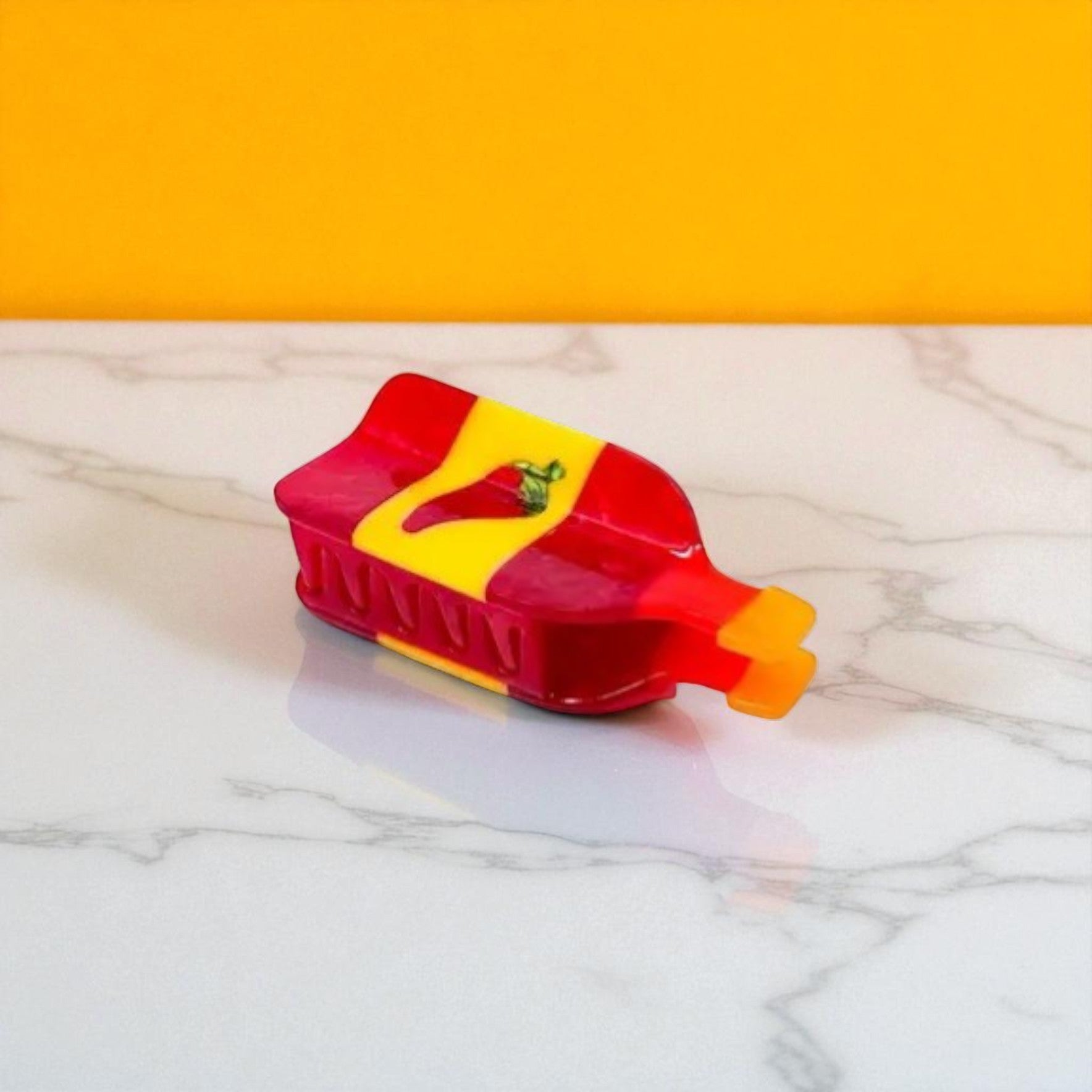 Open red and yellow hot sauce bottle hair clip showing claw teeth, styled on marble with bright yellow backdrop.