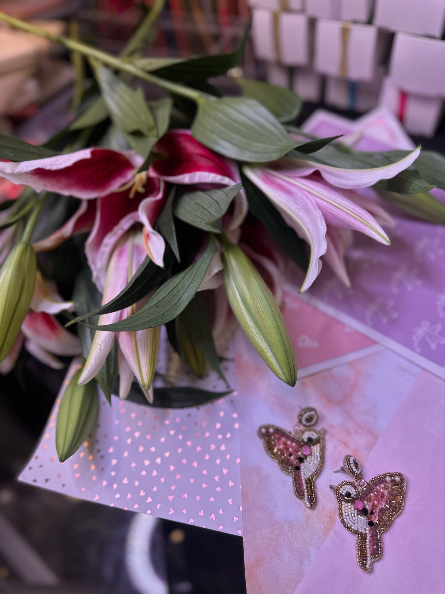 Beaded Magpie Earrings in pink, white, and gold, styled against pastel heart-themed paper with a bouquet of pink lilies in the background, creating a romantic and whimsical display.