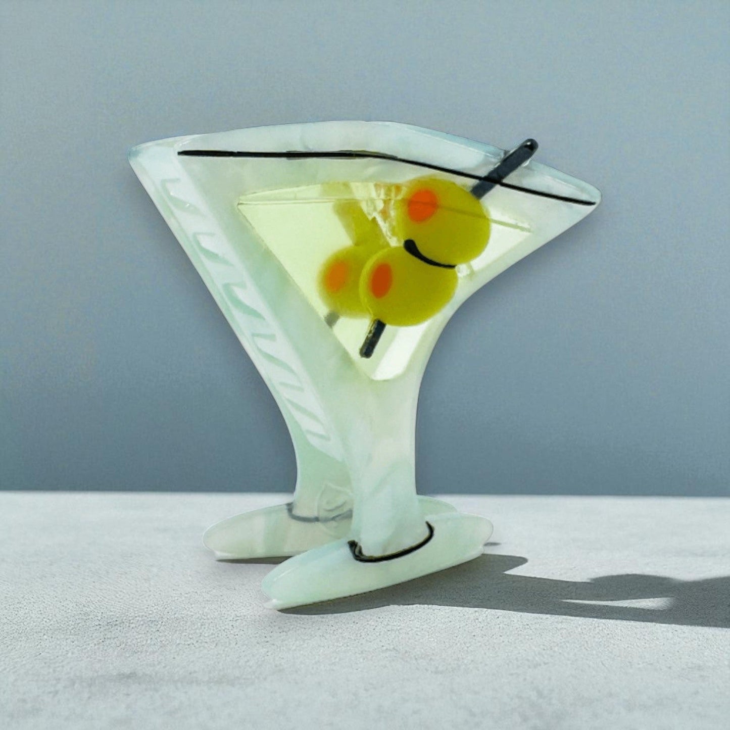 Martini glass claw clip with green olives, angled side view on a grey-blue background.