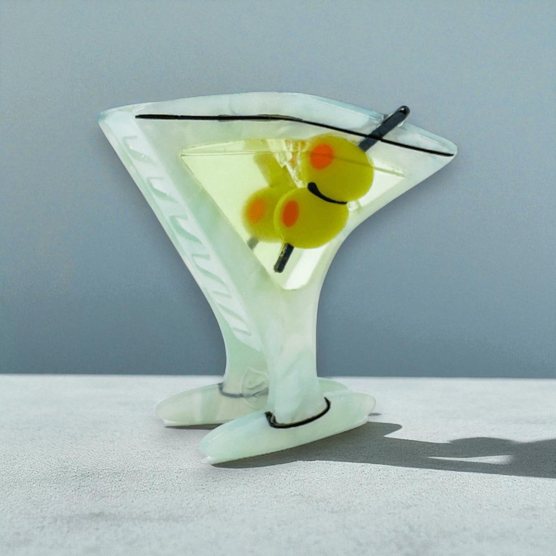 Martini glass claw clip with green olives, angled side view on a grey-blue background.