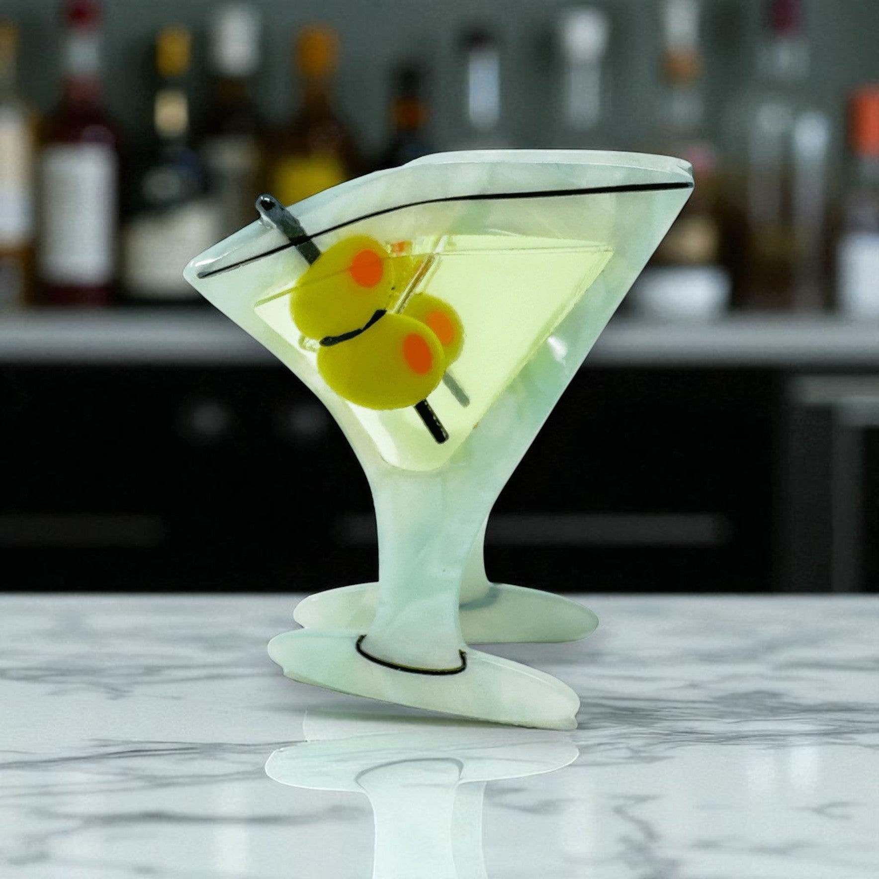 Martini-shaped hair clip with olives standing upright on a marble bar background.