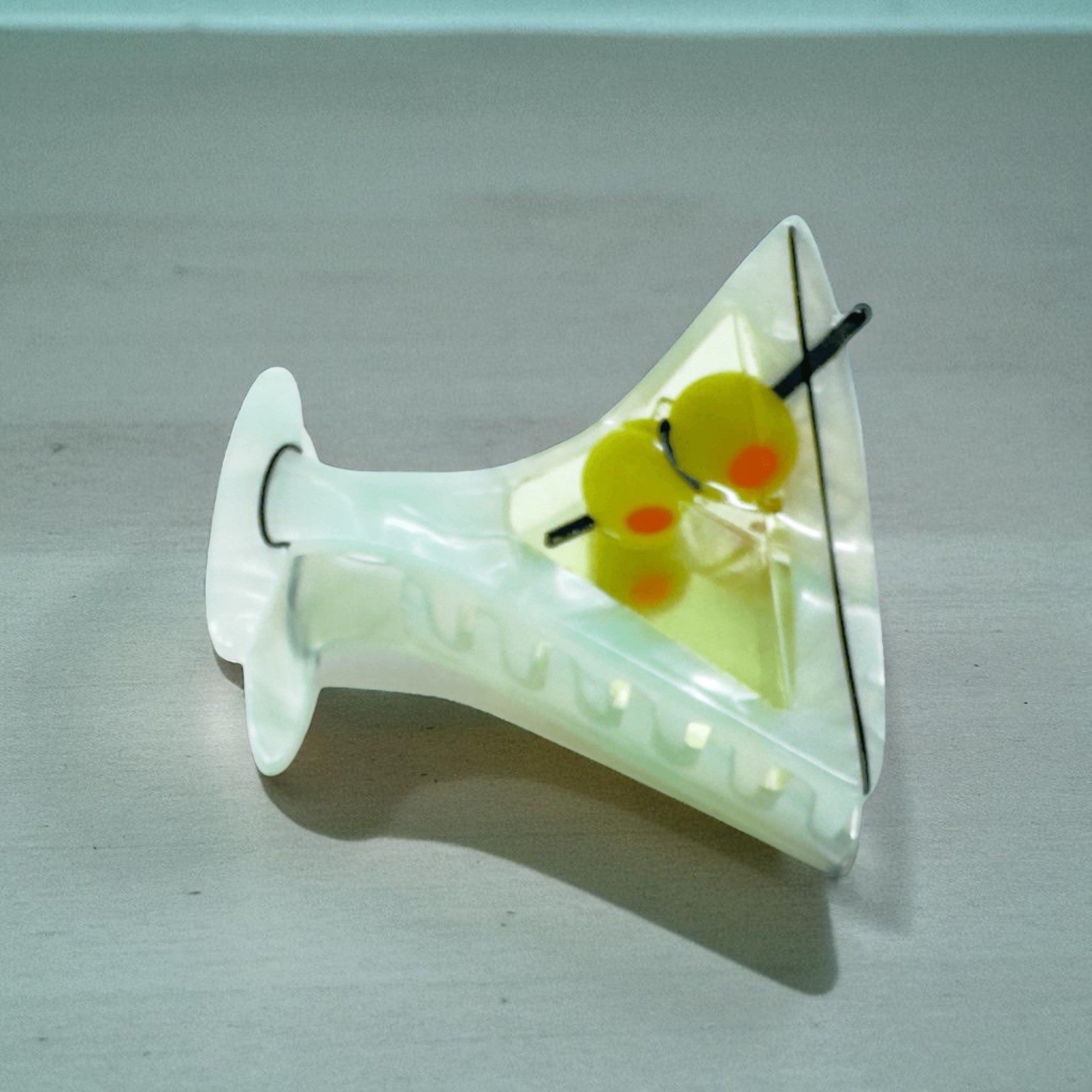 Side flat lay of martini glass hair clip with yellow olives on a light grey surface.
