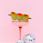 Side view of a green martini olive hair clip perched on a cocktail glass, showing three olive pieces with red pimentos, connected by a skewer.