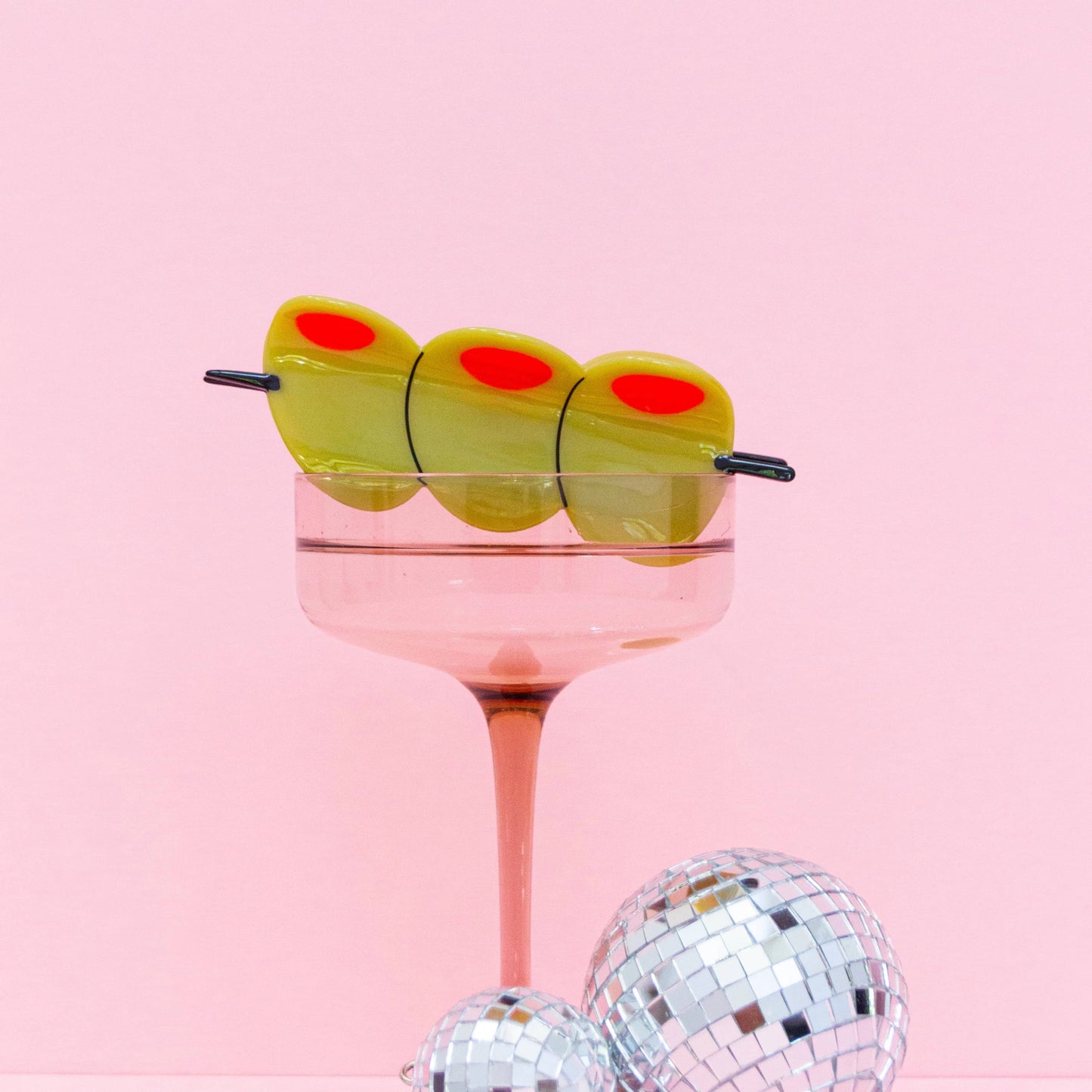 Side view of a green martini olive hair clip perched on a cocktail glass, showing three olive pieces with red pimentos, connected by a skewer.