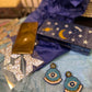Mystical Misfit Blue and Bronze Earrings with evil eye motif displayed on soft tissue paper alongside a celestial-themed clutch and glittering star stickers. Vibrant and whimsical styling for a mystical vibe.