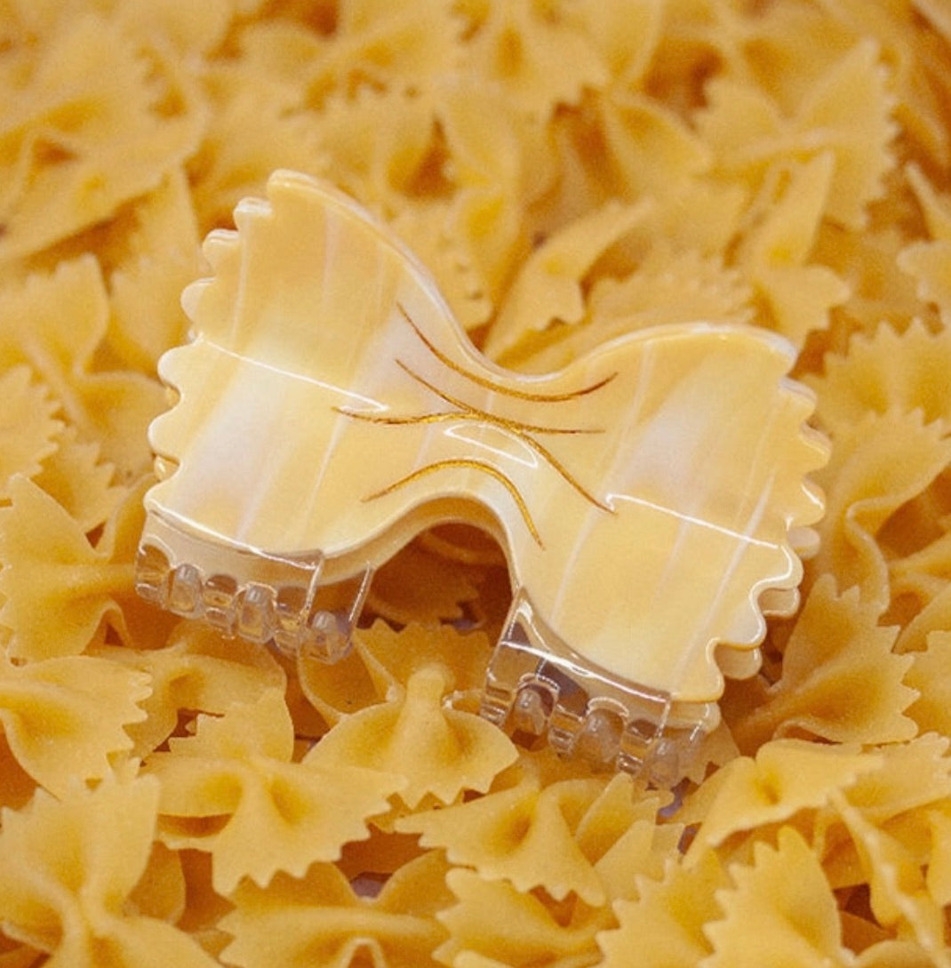 Bowtie pasta-shaped hair claw clip designed to look like farfalle, resting on a bed of real uncooked pasta.
