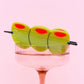 Close-up of a novelty olive skewer hair clip styled on the rim of a pink martini glass with disco balls underneath, set against a pastel pink background.