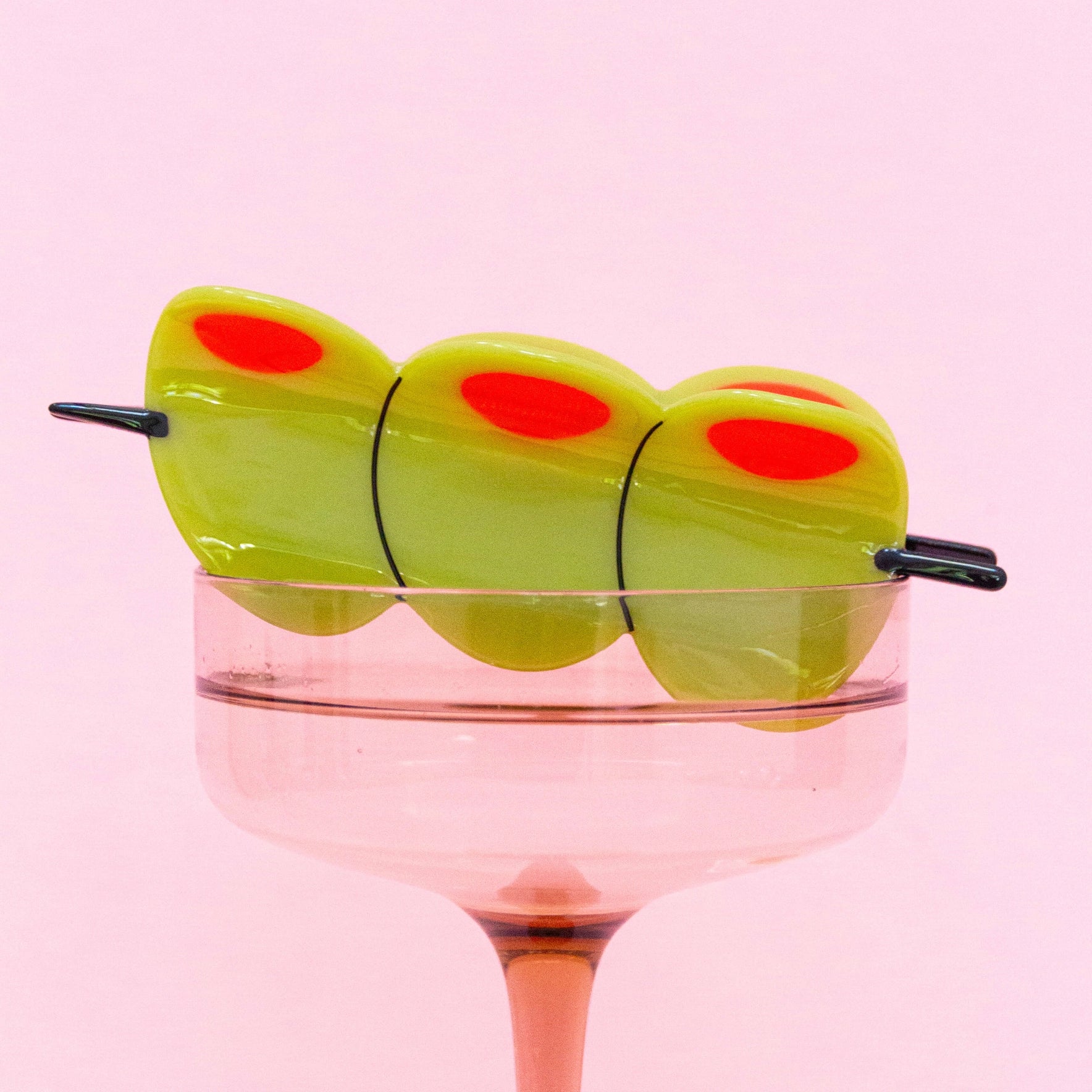 Close-up of a novelty olive skewer hair clip styled on the rim of a pink martini glass with disco balls underneath, set against a pastel pink background.