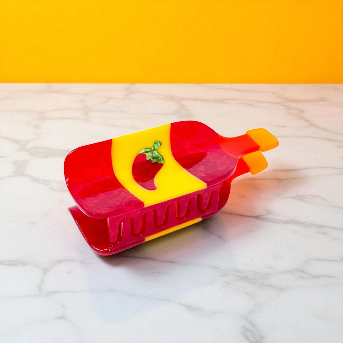 Closed hot sauce-shaped hair clip lying flat on a marble surface with orange background.