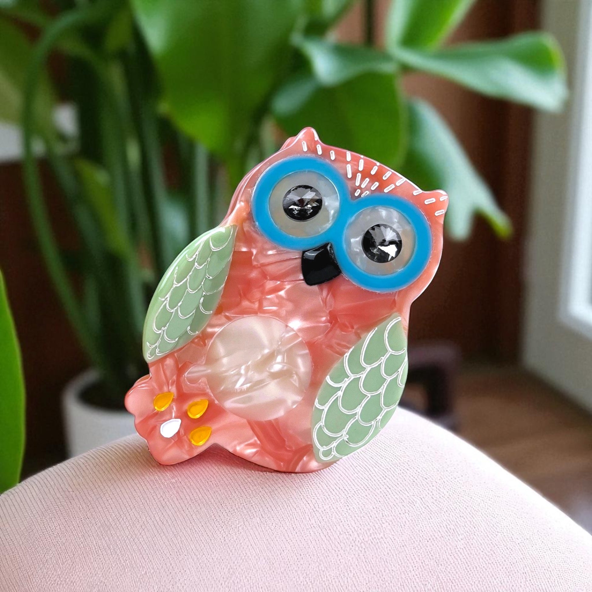 Owl hair clip displayed on a light pink chair with a plant background, showing playful and colorful design.
