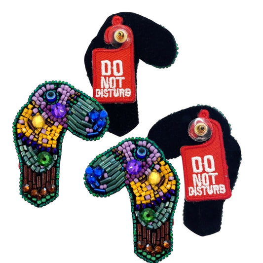Bold statement earrings featuring colorful beaded toucans with vibrant gems and red "Do Not Disturb" embroidered tags on black felt.