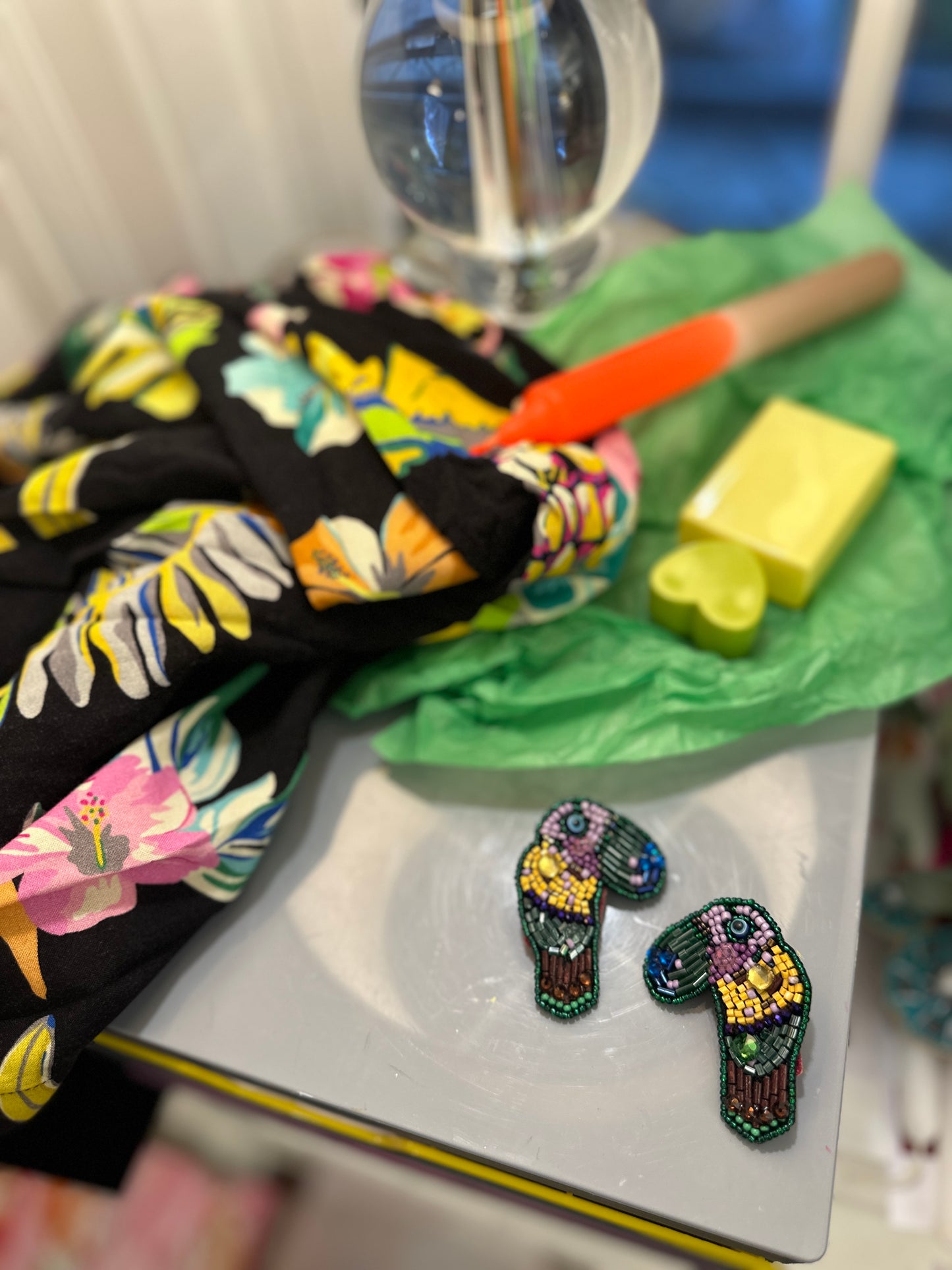 Paradise Plumage Earrings designed as colorful parrots, styled with a vibrant tropical robe, heart-shaped soap, and popsicle decor on a white tabletop.