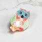 Close-up of a pastel owl-shaped hair clip with big blue glasses, lying on a white marble surface.