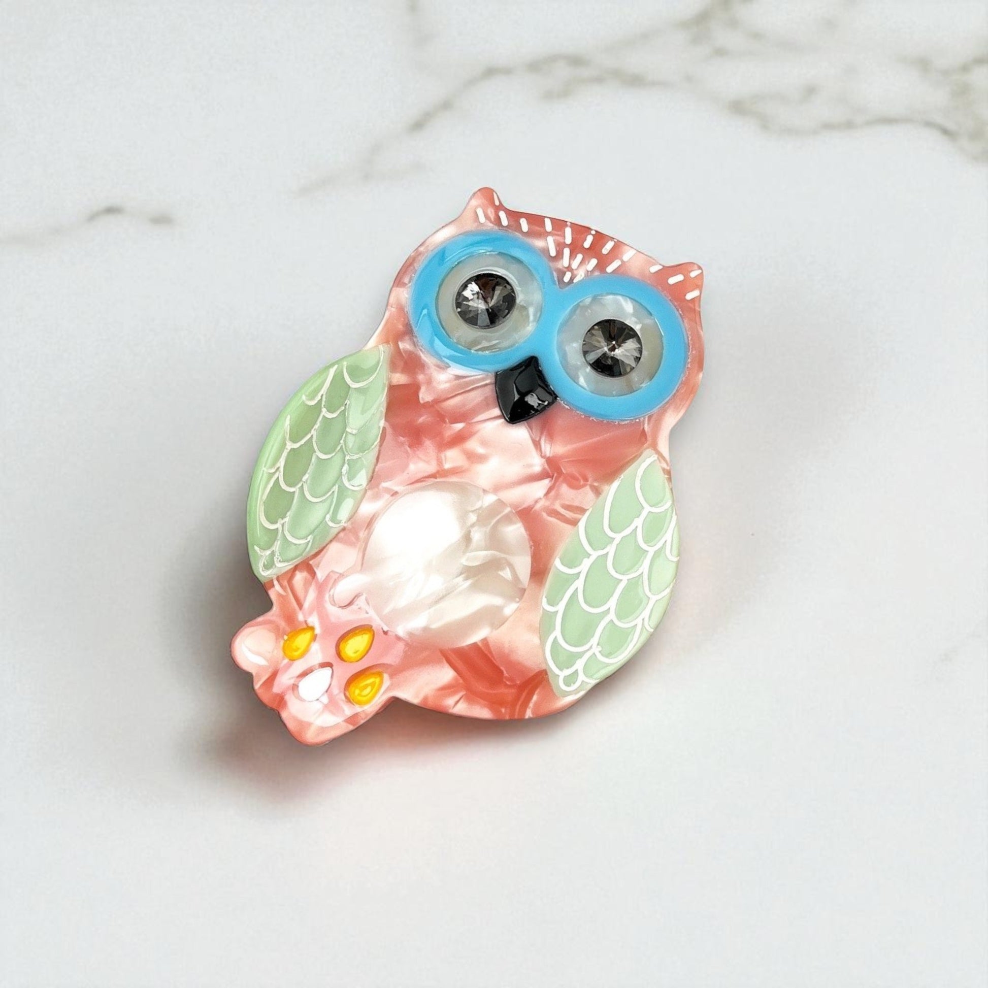 Close-up of a pastel owl-shaped hair clip with big blue glasses, lying on a white marble surface.