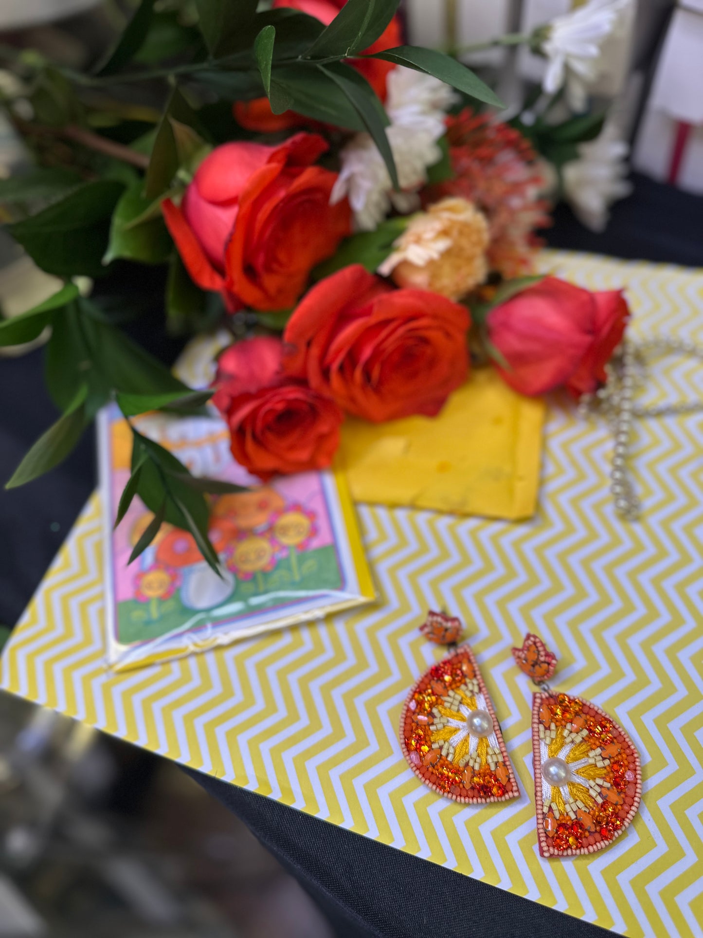 Bright orange citrus-themed earrings styled on a yellow chevron print surface with vibrant red roses, playful flower-themed artwork, and a cheerful aesthetic.
