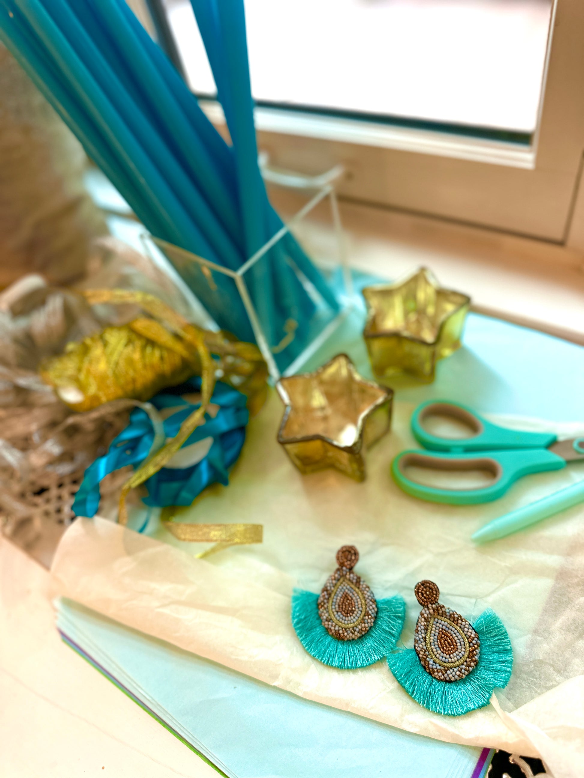 Aqua teardrop fringe earrings with beaded detail, styled with turquoise ribbon, blue straws, scissors, and star-shaped candles for a creative summer vibe.