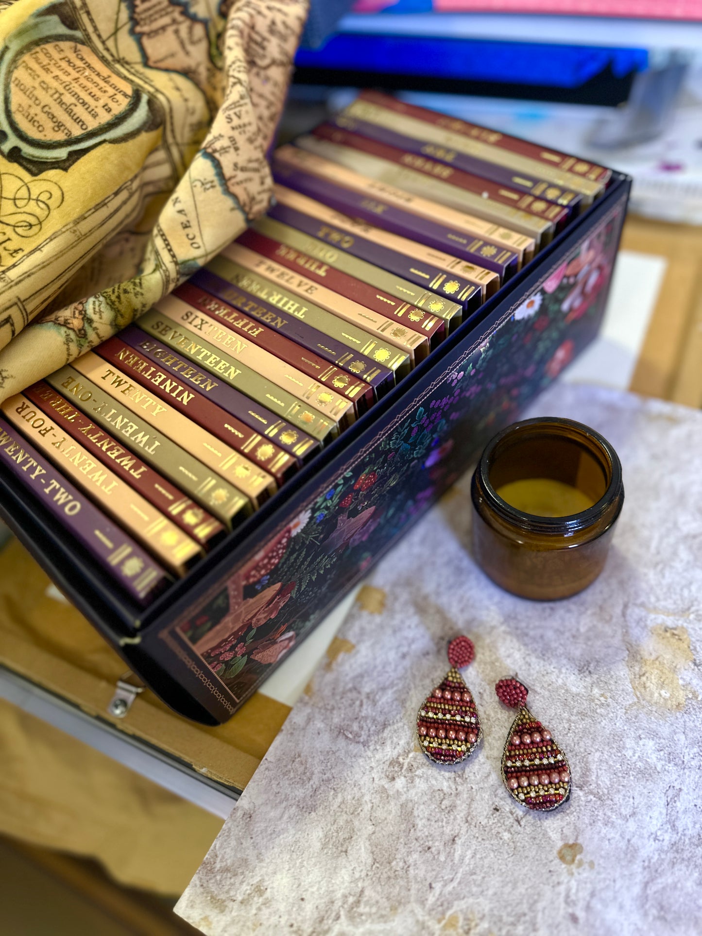 A pair of terra cotta-colored drop earrings featuring intricate beadwork, displayed on a stone surface next to an amber candle jar and a colorful boxed book set.